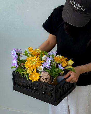 A person in a black shirt and cap holds the Ah!GUGU x Stacked Store Still Life in Bloom - Handcrafted Flowers, featuring vibrant artificial yellow and purple blooms, with a plain light-colored wall as the backdrop.