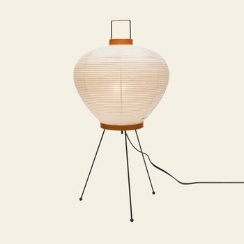 The Akari 3A by Akari is a round white paper lantern lamp with horizontal ribbing, modern design, three slim black metal legs, an orange base, and a black power cord, displayed against a light background.