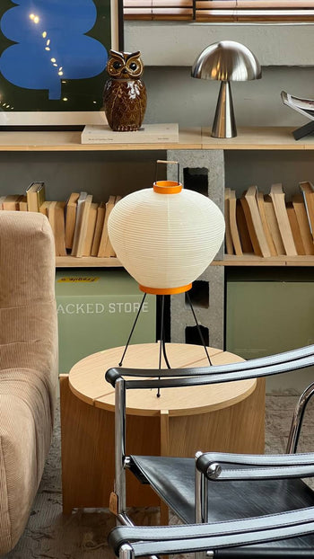 A cozy living room features a round wooden side table with an Akari 3A lamp by Akari, celebrating Japanese craftsmanship. Books are stacked on shelves behind it, with modern chairs enhancing the inviting, modern design.