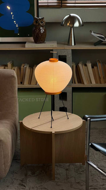 A modern living room features a round wooden side table with the Akari 3A light sculpture by Akari, its glowing paper lantern and black legs showcasing Japanese craftsmanship beside a beige sofa. Stacked books hold a metal owl and mushroom lamp behind.