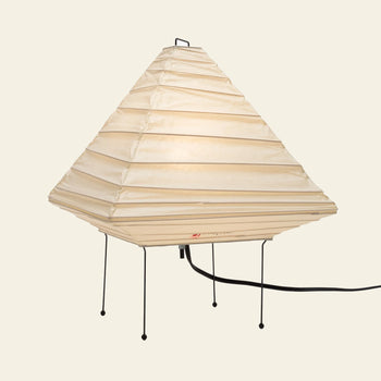 The Akari 5X by Akari is a modern, pyramid-shaped table lamp with a pleated off-white paper shade, slender black metal legs, and a visible black cord, shown against a plain light background.