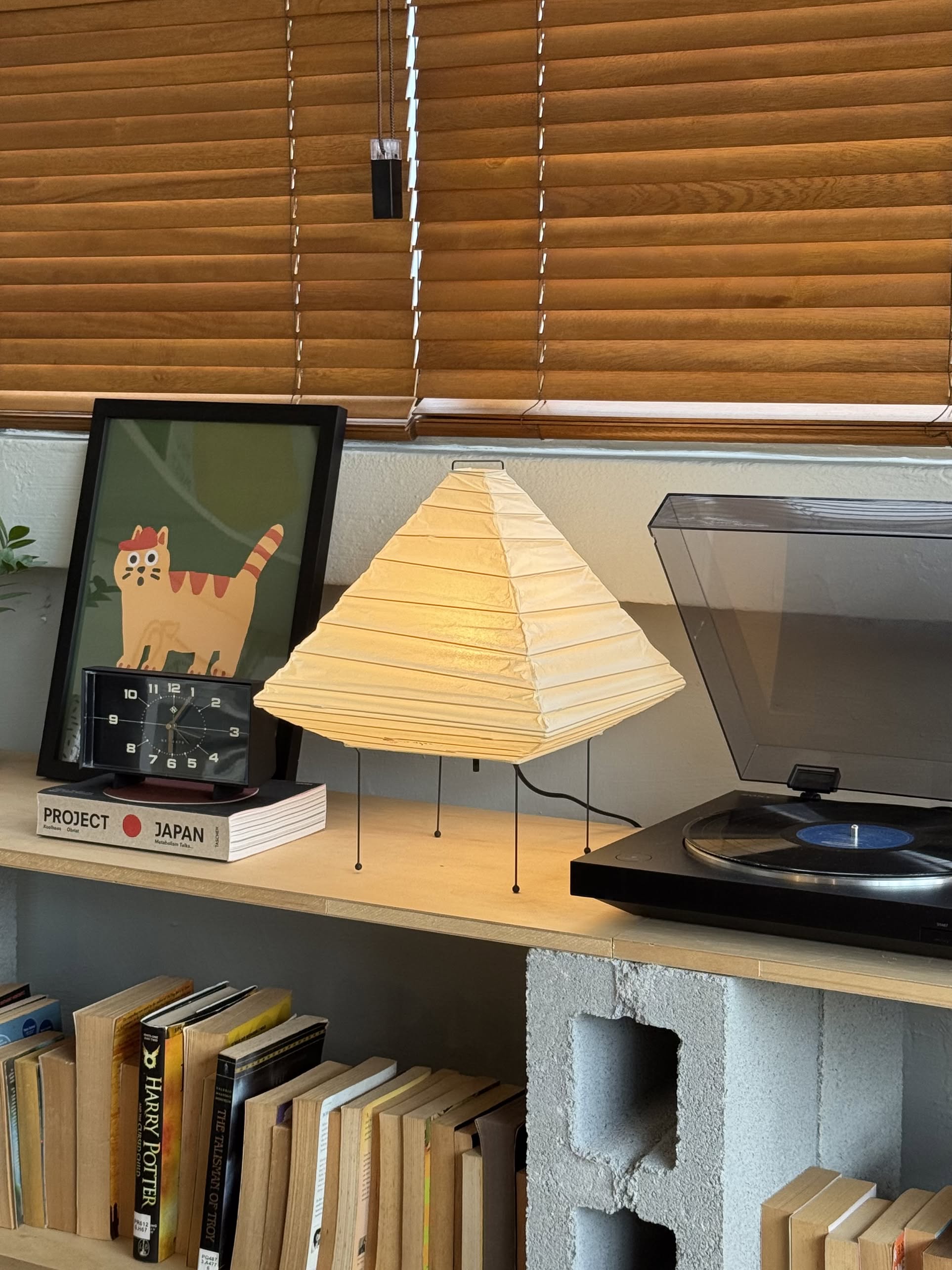 A cozy shelf displays an Akari 5X pyramid-shaped paper lamp by Akari, a framed cat illustration, a flip clock, books, and a record player, all set against wooden blinds.