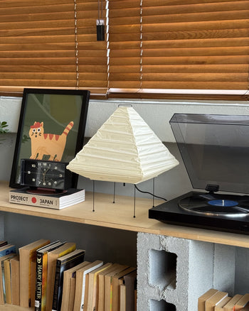 A shelf with books, a framed cat illustration, a retro flip clock, an Akari 5X lamp by Akari bringing modern style, and a record player with vinyl, all beneath wooden blinds.
