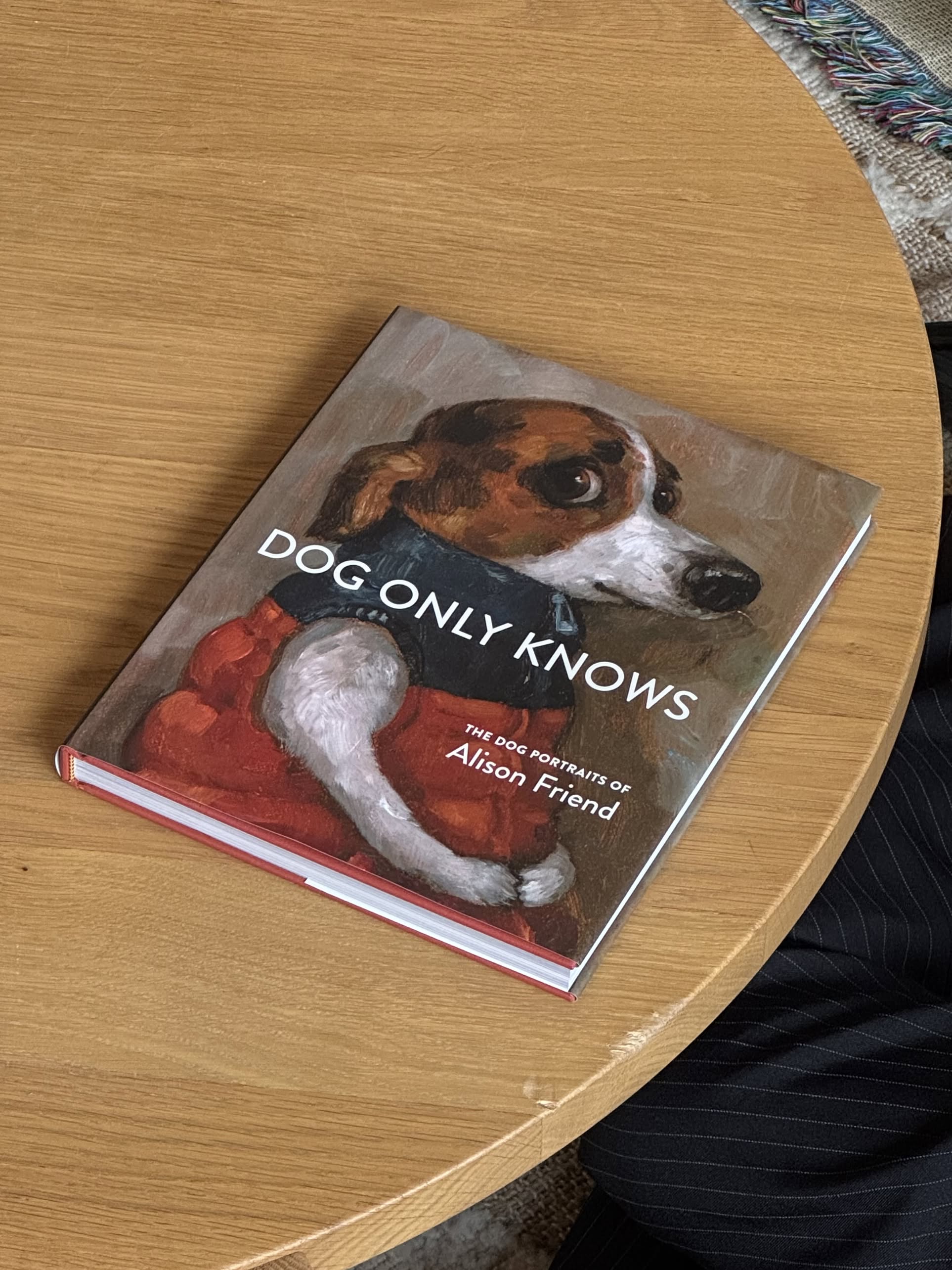 Alison Friend - Dog Only Knows by Alison Friend sits on a wooden table, its cover displaying delightful oil-painted dog art—a pup in a red vest alongside part of a person’s leg. Perfect for dog lovers and art enthusiasts alike.