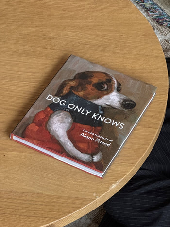 Alison Friend - Dog Only Knows by Alison Friend sits on a wooden table, its cover displaying delightful oil-painted dog art—a pup in a red vest alongside part of a person’s leg. Perfect for dog lovers and art enthusiasts alike.