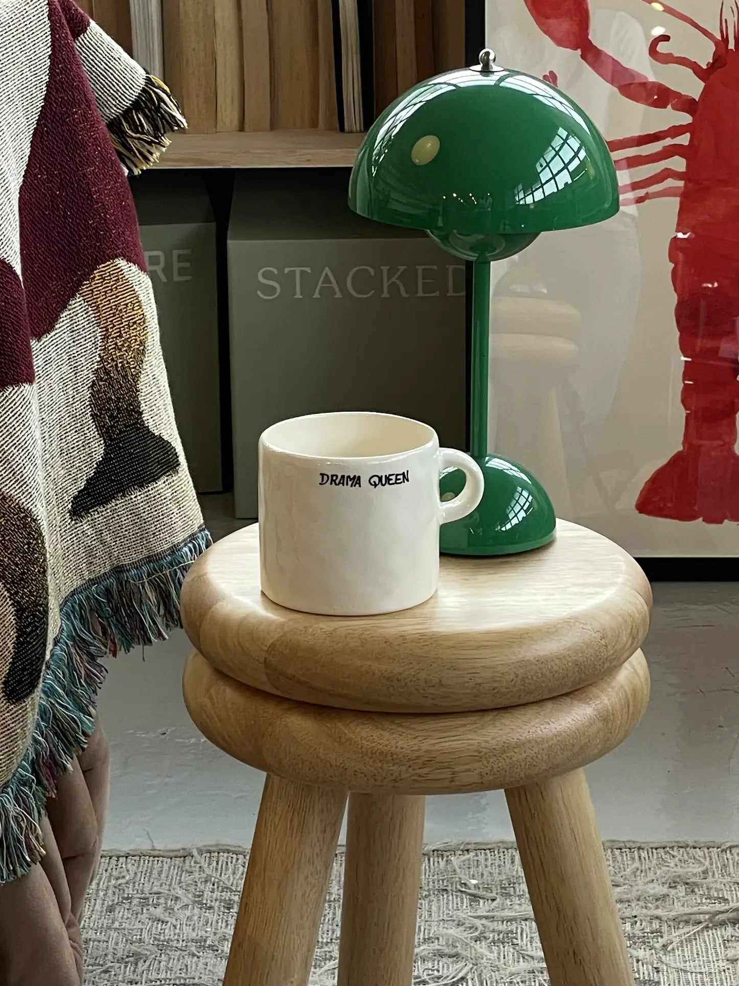 The Anna + Nina Drama Queen Mug by Anna + Nina sits on a wooden stool next to a green table lamp, bringing theatrical flair to your coffee breaks, while a fringed blanket and artwork enhance the expressive, cozy vibe.