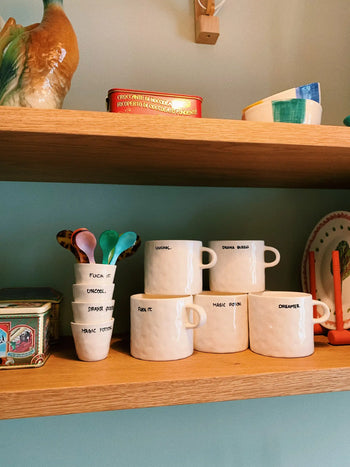 The Anna + Nina Drama Queen Mug, featuring a handwritten label, brings theatrical charm to wooden shelves. Paired with colorful spoons and vintage tins, it creates a decorative setting ideal for lively coffee breaks.