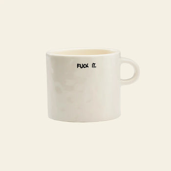 The Anna + Nina Fuck It Mug by Anna + Nina is a plain white ceramic mug with a handle, featuring “FUCK IT.” in bold black uppercase letters on one side—perfect for adding attitude to your coffee or tea break. Set against a light beige background.