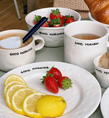 A breakfast table with Anna + Nina Good Morning Bowls, a cappuccino-filled mug, tea, fresh strawberries, lemon slices and a whole lemon, plus a croissant. A blue pen rests on one mug.