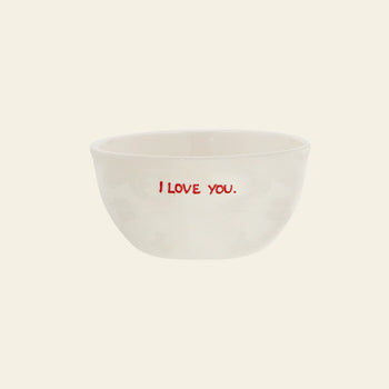 The Anna + Nina I Love You Bowl is a small white ceramic bowl from Anna + Nina, featuring I LOVE YOU. in red on the front—perfect for breakfast and displayed against a plain, light background.