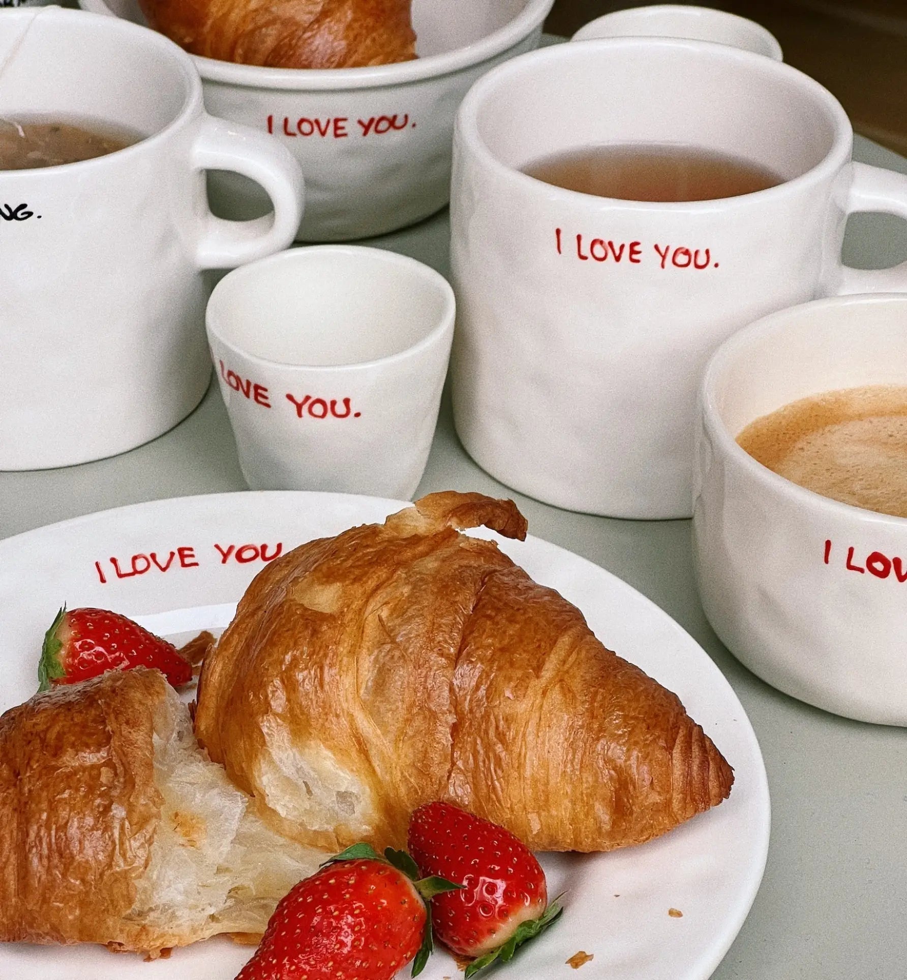 A cozy breakfast scene features croissants and strawberries served with coffee or tea in the Anna + Nina I Love You Bowl, all white with bold red “I LOVE YOU.” text, creating an inviting start to the day.