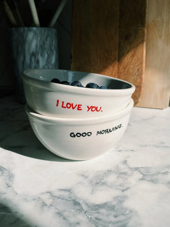 Two Anna + Nina I Love You Bowls are stacked on a marble countertop; the top bowl, filled with blueberries, features I LOVE YOU. in red, while the bottom has GOOD MORNING. in black—a charming breakfast set from Anna + Nina.