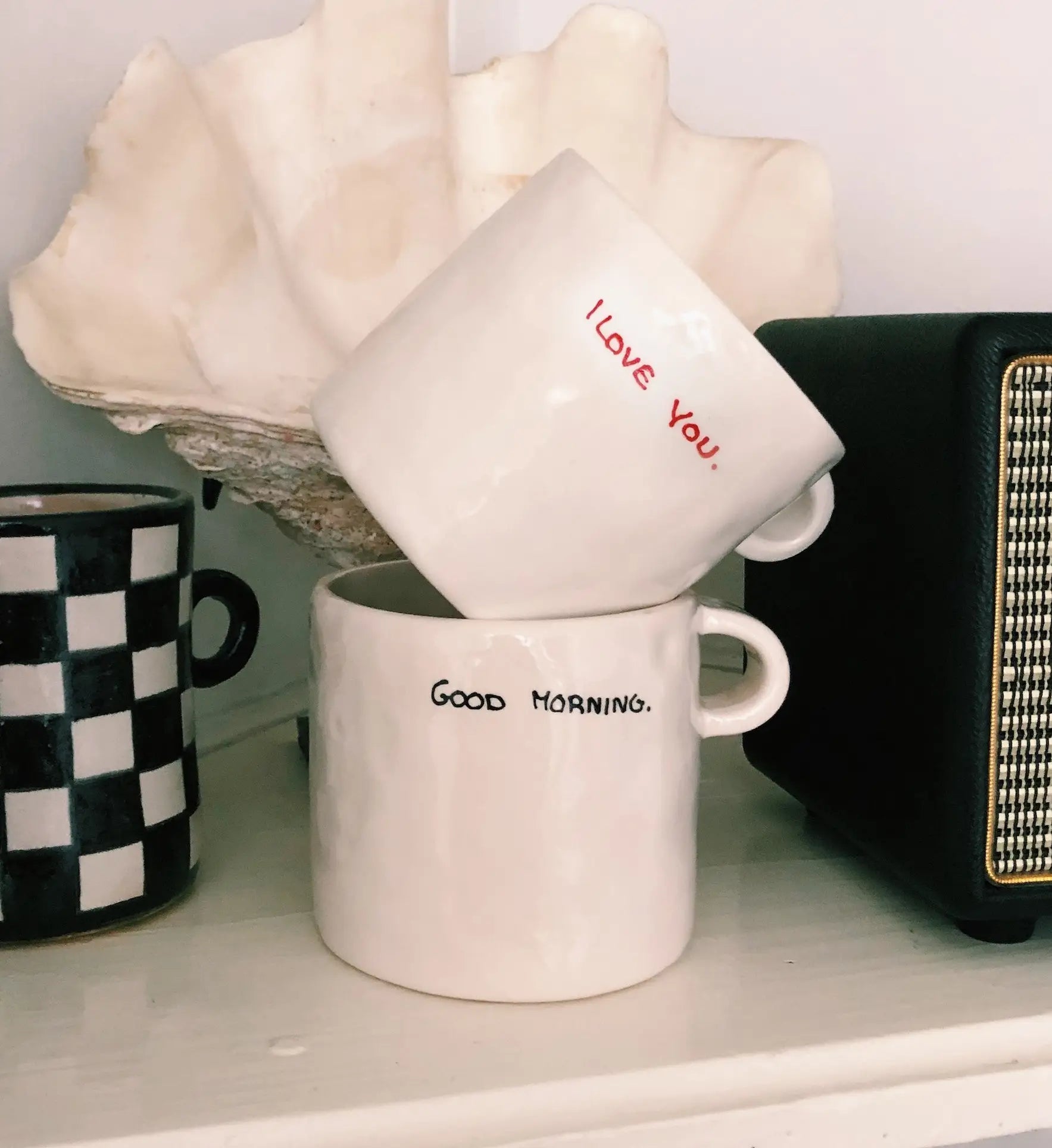 Two white mugs are stacked on a shelf; the top is the Anna + Nina I Love You Mug, ideal for expressing affection. Nearby are a seashell, coffee in a black-and-white checkered mug, and a black speaker.