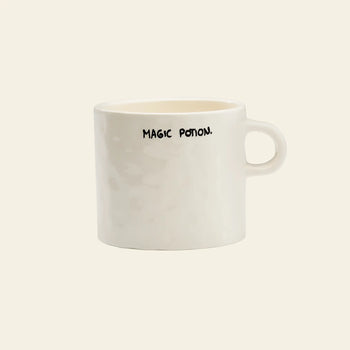 The Anna + Nina Magic Potion Mug is a plain white mug by Anna + Nina, featuring MAGIC POTION. in black uppercase letters near the rim, and is perfect for your next brew against a simple, light background.