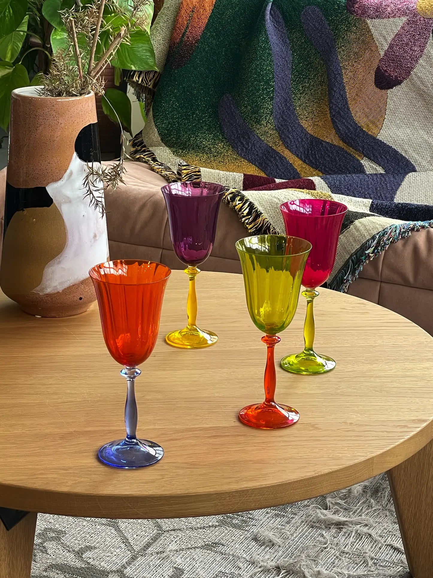 The Anna + Nina Multicoloured Wine Glasses, Set of 4, are displayed on a light wooden table alongside a ceramic vase with dried flowers and a patterned blanket, creating a cozy vintage-inspired scene.