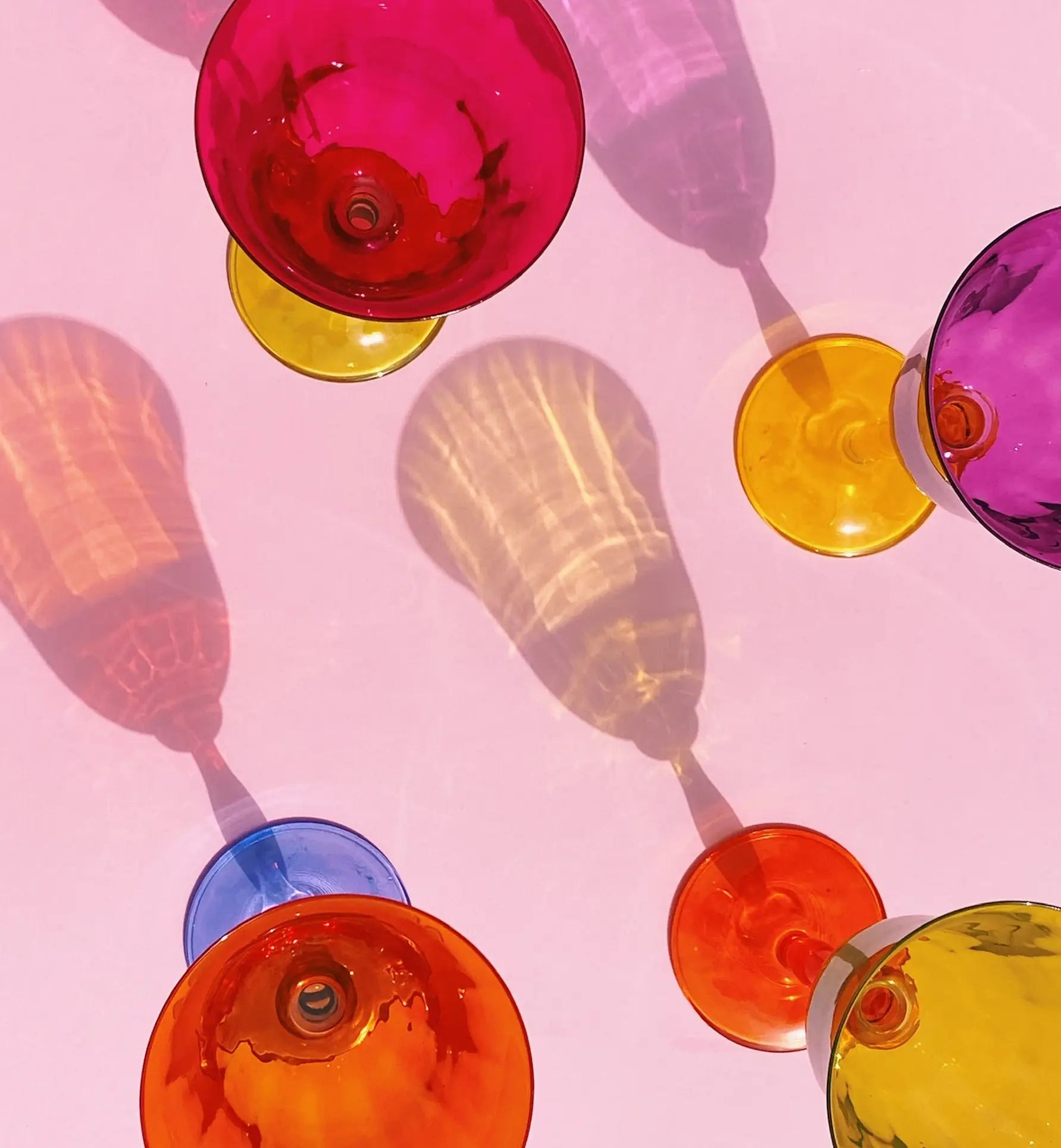 The Anna + Nina Multicoloured Wine Glasses, Set of 4, feature vibrant bases in red, orange, yellow, and blue. Their playful vintage design casts lively shadows—an ideal housewarming gift from Anna + Nina.