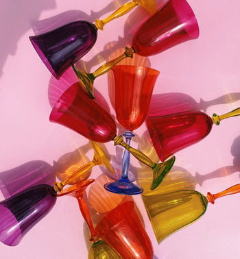 The Anna + Nina Multicoloured Wine Glasses, Set of 4, features vibrant red, yellow, purple, and blue shades on a pink surface—playfully arranged to create vintage charm. Perfect as a housewarming gift or artistic centerpiece.
