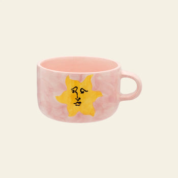 The Anna + Nina Sunny Side Up Cappuccino Mug features a pink marbled texture, a yellow sun with a stylized face, and a simple handle—perfect for brightening your coffee moments.