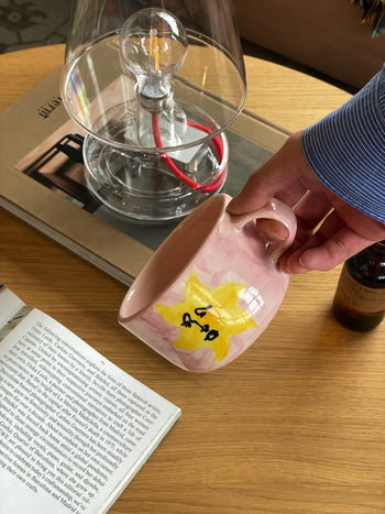 A hand holding the Anna + Nina Sunny Side Up Cappuccino Mug, featuring a yellow star and black writing, above a wooden table with a book, magazine, clear lamp, and bottle—perfect for a cozy coffee moment.