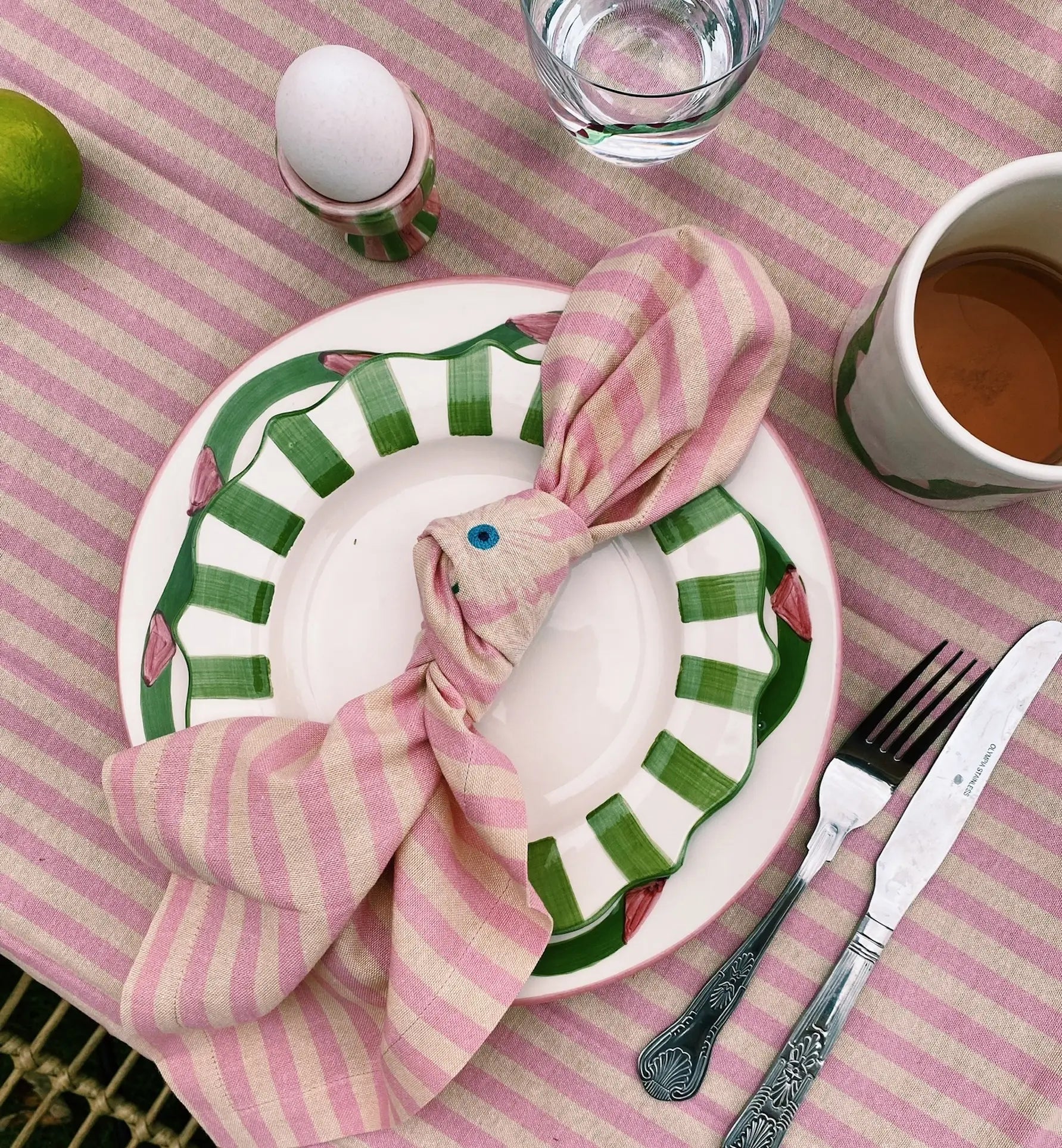 A table set with a green and white plate, Anna + Nina Trippy Flower Napkin folded like a fish with a blue eye, pink-striped tablecloth, glass of water, cup of tea, lime, and a soft-boiled egg for a spring floral vibe.