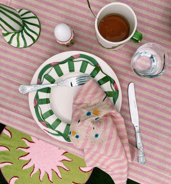 A striped pink and white tablecloth sets a spring vibe with plates, cutlery, glassware, and an Anna + Nina Trippy Flower Napkin, known for its playful stripes and bold blue and yellow dots, folded neatly on the plate.