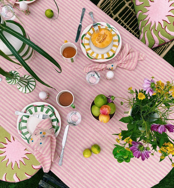 A colorful table set for a meal with a pink striped tablecloth, green and white plates, sliced melon, cups of tea, flowers, fruit bowl, and Anna + Nina Trippy Flower Napkins (Set of 2) by Anna + Nina adds to the floral spring vibe.