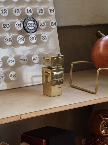The Anna + Nina Back to the Future Figure, a small brass piece, is placed on a wooden shelf next to a white perpetual calendar and other decor. Behind it are a brown object and part of a gold-finished metal frame.