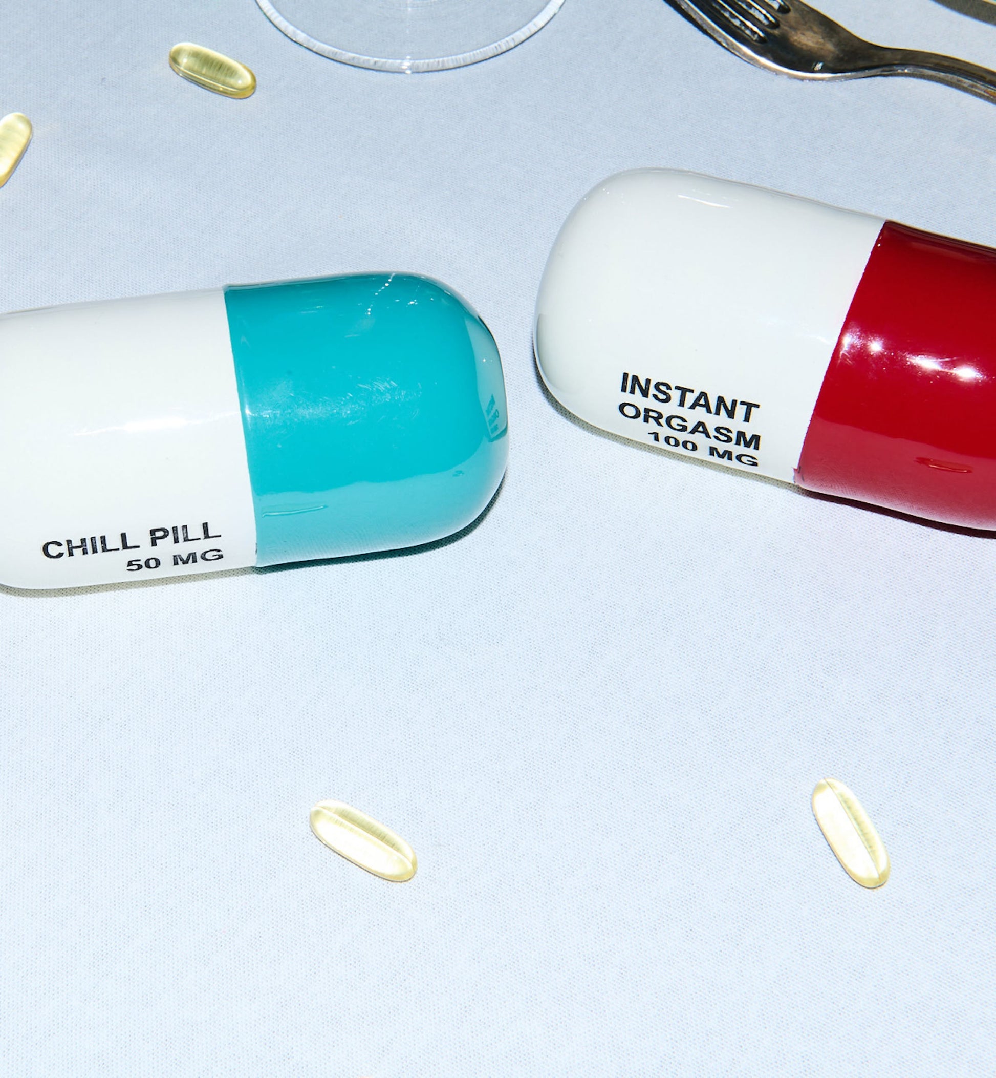 The Anna + Nina Chill Pill Figure features two large capsules—one labeled INSTANT ORGASM 100 MG—surrounded by small yellow capsules. The multicoloured design pops on a white surface, with a fork partially visible in the scene.