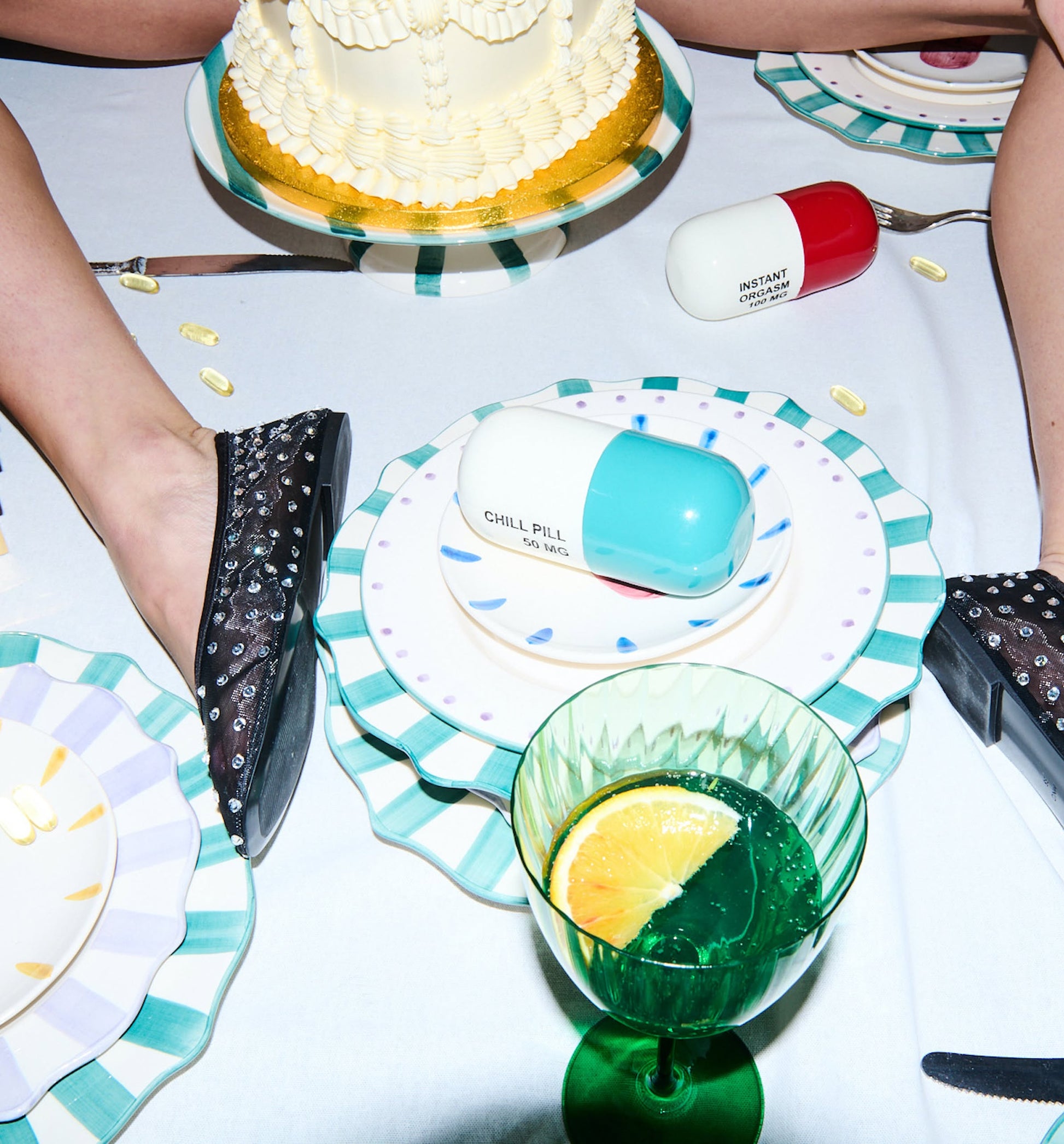A playful table setting showcases the Anna + Nina Chill Pill Figure containers in a multicoloured design, alongside a cake, decorative plates, a green glass with lemon slice, scattered capsules, and two feet in sparkly shoes resting on the table.