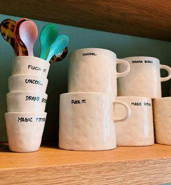 A wooden shelf displays Anna + Nina ceramic mugs and cups, including the Drama Queen Espresso Cup with bold phrases like FUCK IT., UNCOOL., and MAGIC POTION. Four colorful spoons complete this playful, luxurious set.
