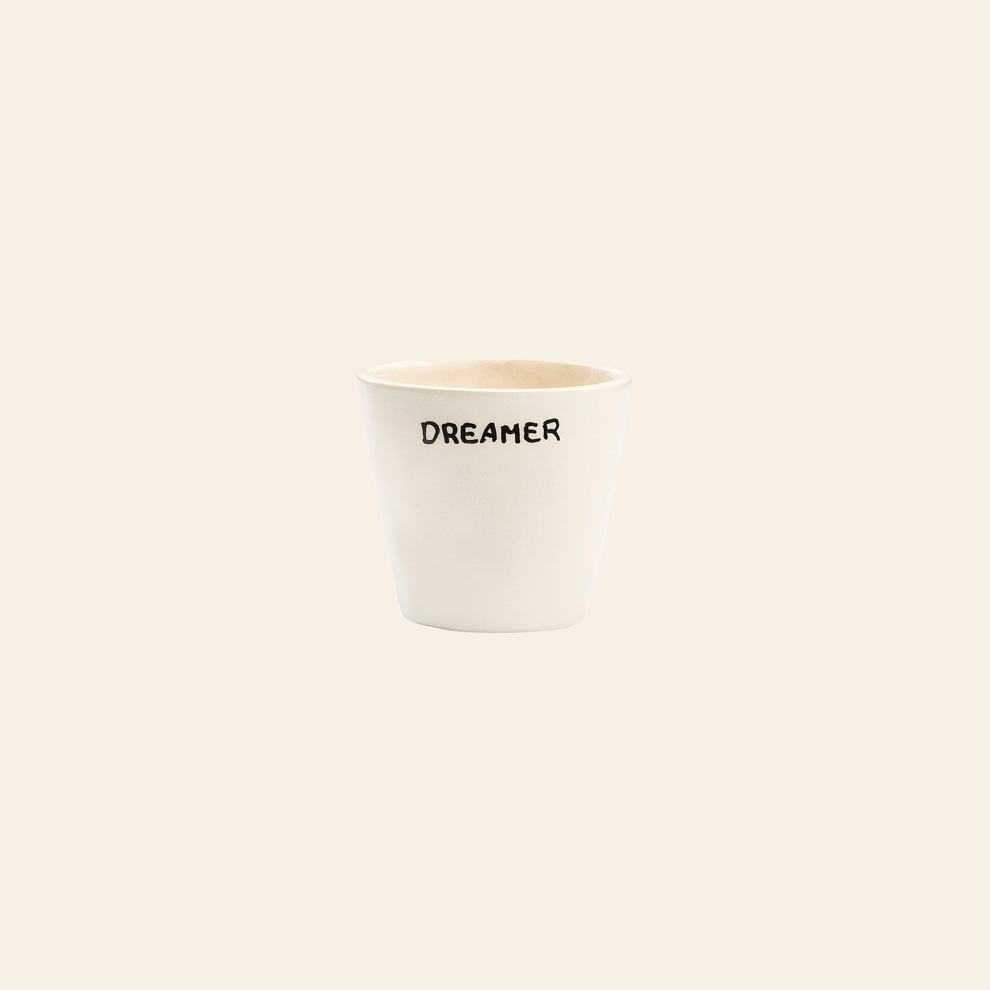 The Anna + Nina Dreamer Espresso Cup is a handcrafted white ceramic cup featuring the word DREAMER in black, centered on a plain beige background—a minimalist and elegant design by Anna + Nina.