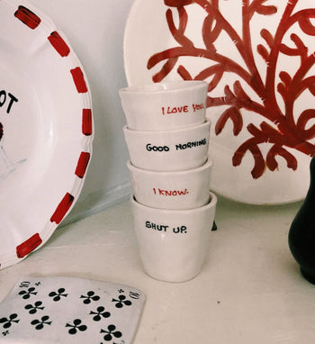 Four Anna + Nina I Love You Espresso Cups in white are stacked, each featuring a unique phrase. Decorative plates and a playing card appear in the background—ideal for gifting or adding to your collection.