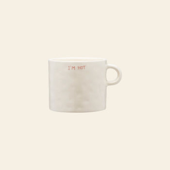 The Anna + Nina Im Hot Mug is a plain white ceramic mug with a handle, featuring IM HOT in small red letters on the front—ideal for hot drinks or gifting. Shown against a light beige background.
