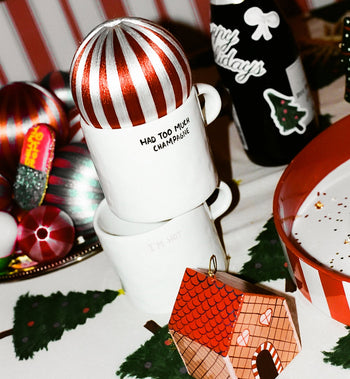 Two stacked Anna + Nina Im Hot Mugs sit beside hot drinks and a black Happy Holidays bottle, all surrounded by holiday décor—making this festive Anna + Nina setup perfect for gifting or cozy celebrations.