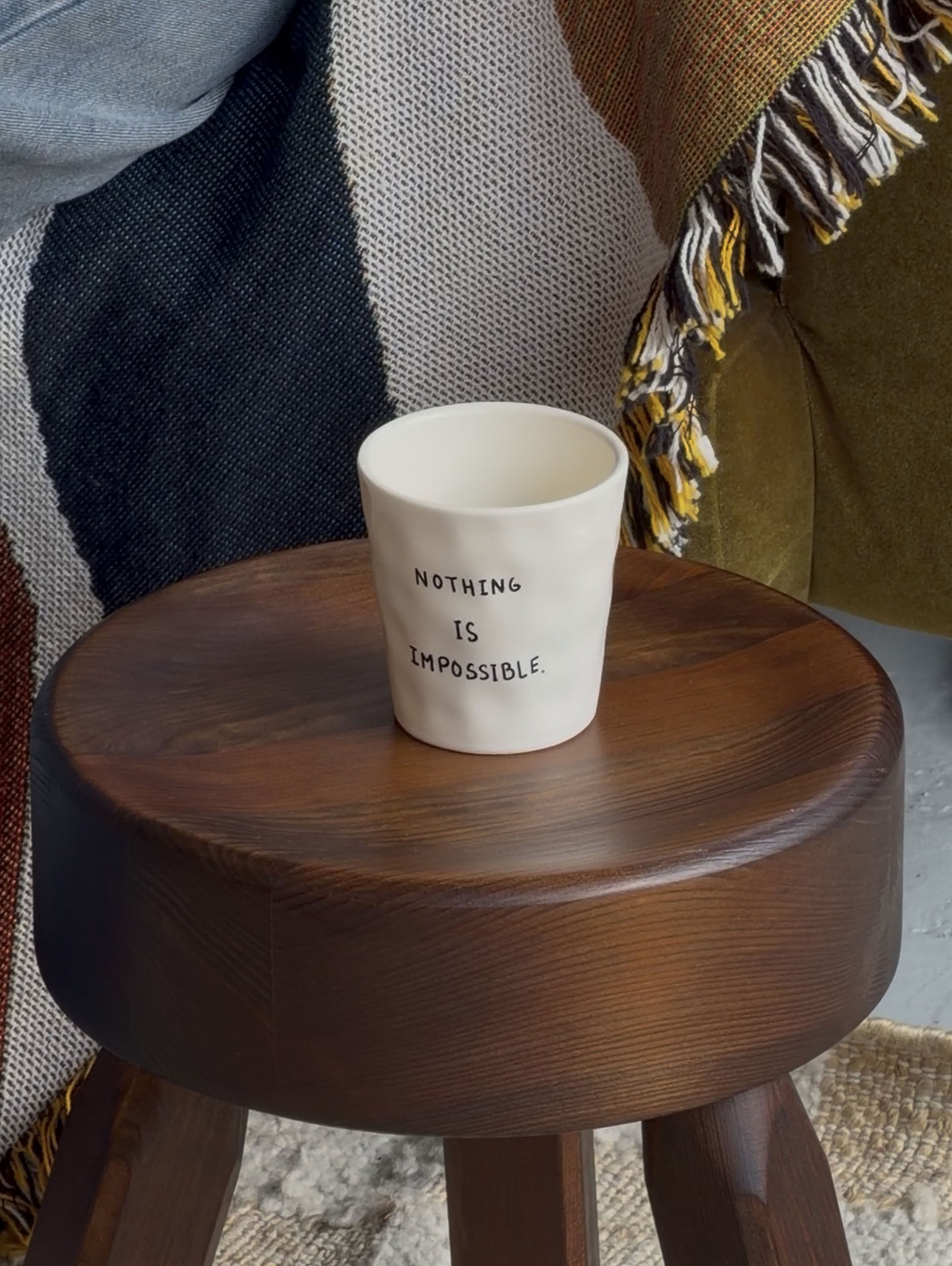 The Anna + Nina Nothing Is Impossible Coffee Cup by Anna + Nina sits on a dark wooden stool beside a textured blanket draped over a green chair.