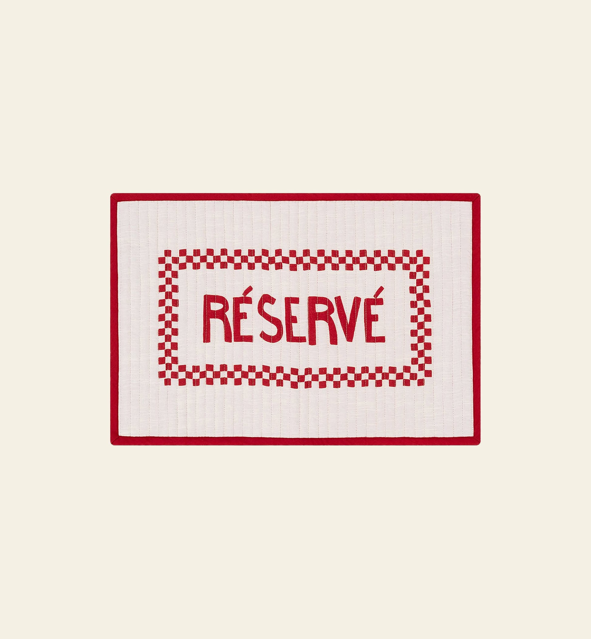 The Anna + Nina Reserve Placemat by Anna + Nina features a rectangular white design with a red border, checkered pattern, and bold RÉSERVÉ at the center. Made from organic cotton, it adds French flair when displayed on a light beige surface.