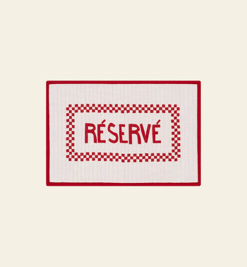 The Anna + Nina Reserve Placemat by Anna + Nina features a rectangular white design with a red border, checkered pattern, and bold RÉSERVÉ at the center. Made from organic cotton, it adds French flair when displayed on a light beige surface.