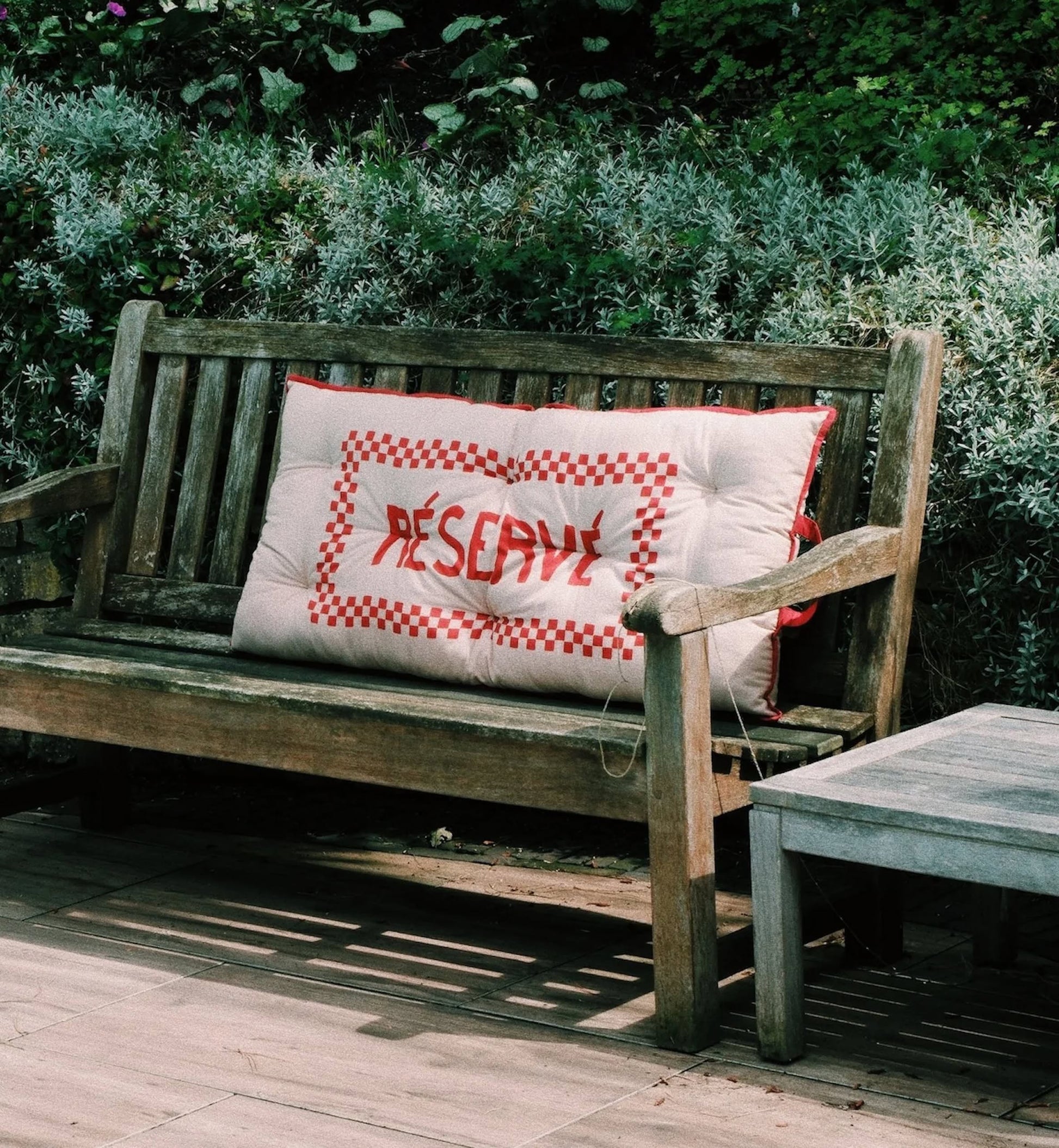 A wooden garden bench with a large cushion featuring red checkered edges and RECEPTION stitched in red evokes French flair, reminiscent of the Anna + Nina Reserve Placemat, set amid lush green and gray foliage outdoors.