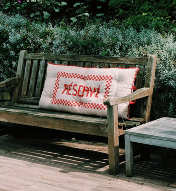 A wooden garden bench with a large cushion featuring red checkered edges and RECEPTION stitched in red evokes French flair, reminiscent of the Anna + Nina Reserve Placemat, set amid lush green and gray foliage outdoors.