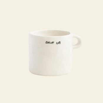 The Anna + Nina Shut Up Mug is a white ceramic mug with a small handle and bold black SHUT UP. text on the side—ideal for your daily caffeine fix.