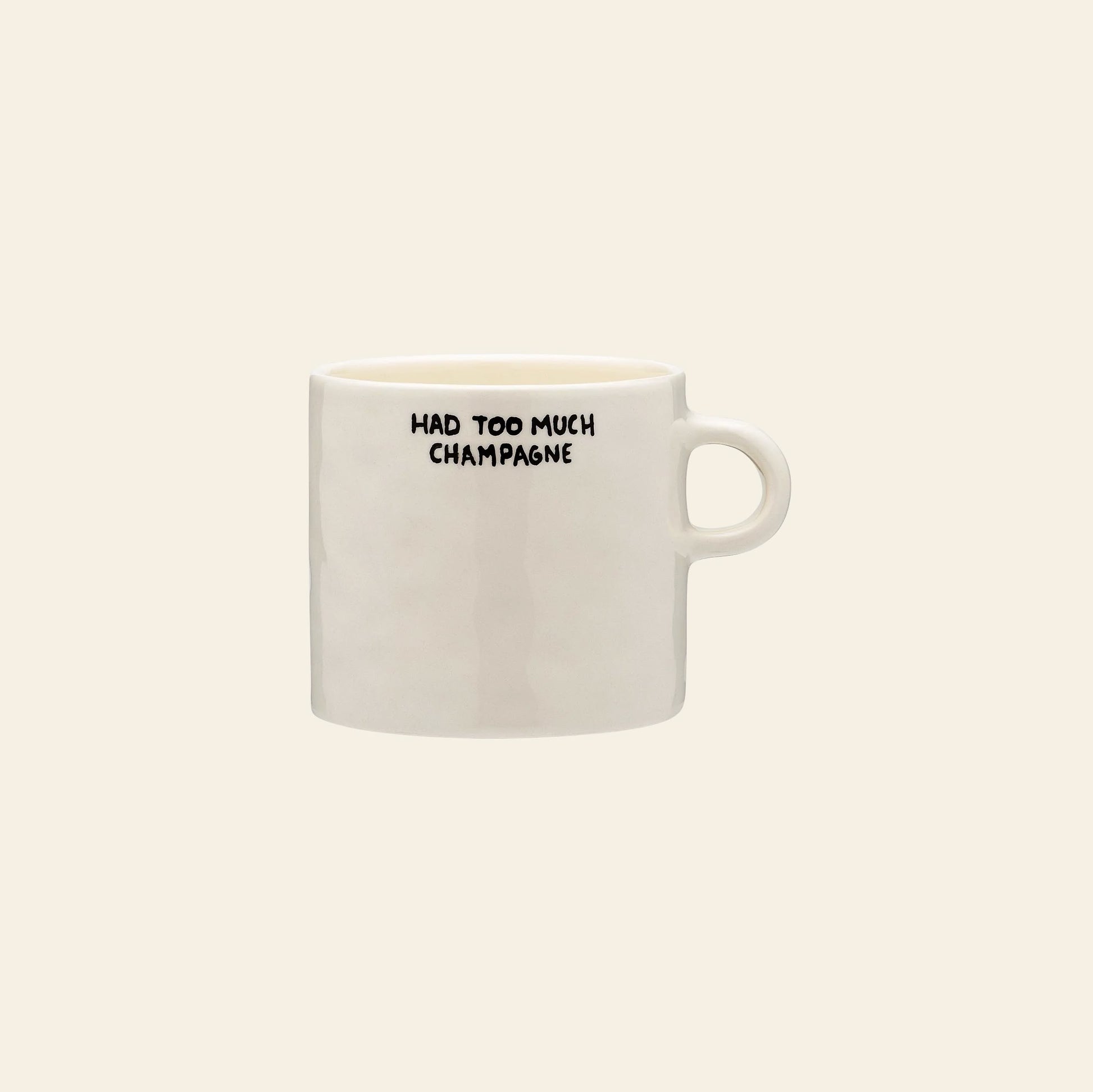 The Anna + Nina Too Much Champagne Mug by Anna + Nina is a white ceramic mug featuring HAD TOO MUCH CHAMPAGNE in bold black letters near the rim, displayed against a beige background.