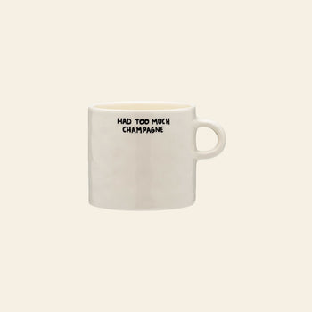 The Anna + Nina Too Much Champagne Mug by Anna + Nina is a white ceramic mug featuring HAD TOO MUCH CHAMPAGNE in bold black letters near the rim, displayed against a beige background.