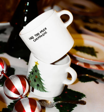 Two white ceramic mugs are stacked; the top is the Anna + Nina Too Much Champagne Mug by Anna + Nina, ideal for champagne lovers, while the bottom displays a Christmas tree design. Decorative ornaments and festive tableware surround them.