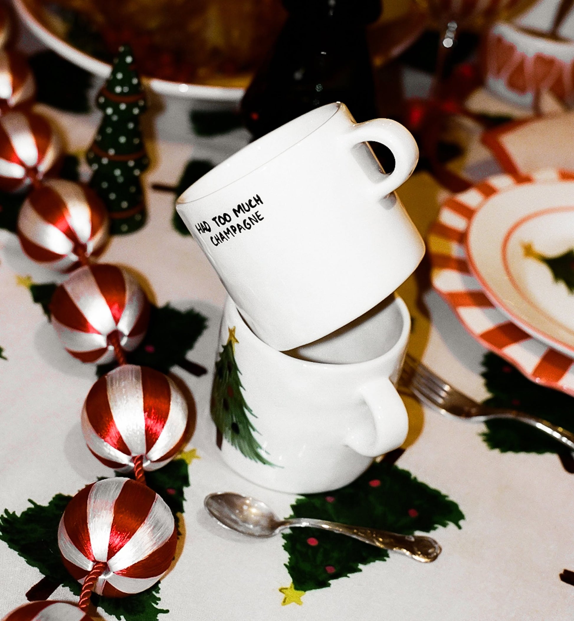 Two white Anna + Nina ceramic mugs are stacked on a festive table with Christmas decorations. The top mug reads HAD TOO MUCH CHAMPAGNE—a perfect Anna + Nina Too Much Champagne Mug for champagne lovers.