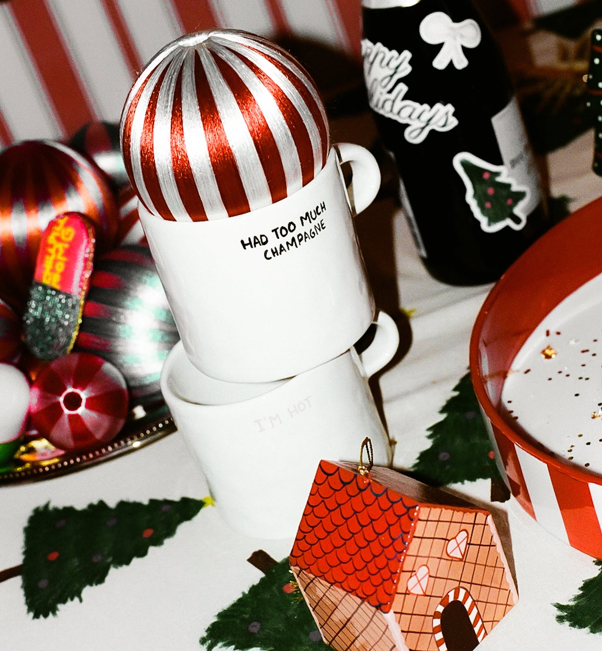 Two stacked Anna + Nina Too Much Champagne Mugs rest on a festive table with ornaments, a bottle, a striped plate, and a decorative house—an ideal holiday gift for any champagne lover.