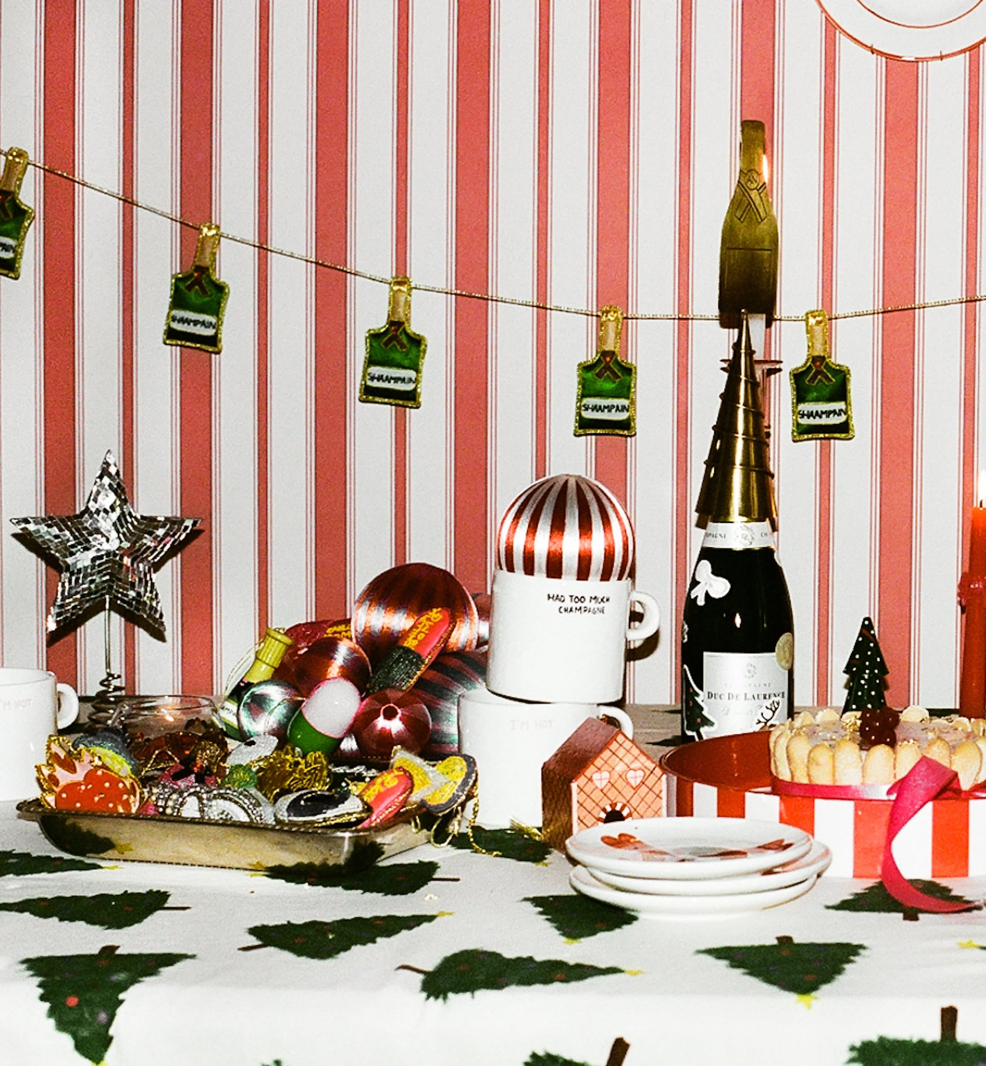 A festive table is adorned with Christmas decorations, cookies, ornaments, a striped centerpiece, and the Anna + Nina Too Much Champagne Mug by Anna + Nina, set against red and white striped wallpaper with hanging green and gold tags.