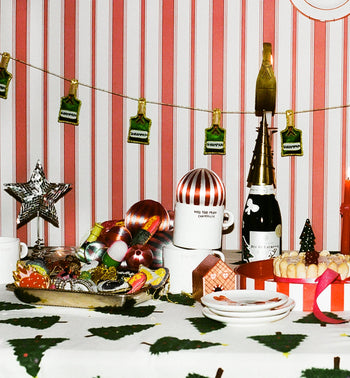 A festive table is adorned with Christmas decorations, cookies, ornaments, a striped centerpiece, and the Anna + Nina Too Much Champagne Mug by Anna + Nina, set against red and white striped wallpaper with hanging green and gold tags.