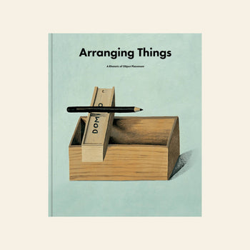 The cover of Apartamento Arranging Things: A Rhetoric of Object Placement by Apartamento shows a wooden box with dominoes and a pencil on top, reflecting its design ethos against a soft, light background.