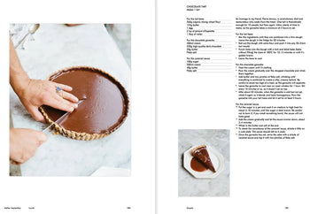 A person cuts a chocolate tart in a fluted pan. On the right is a recipe from Apartamento Atelier September: A Place for Daytime Cooking by Apartamento, with a photo of one tart slice on a plate next to a fork.