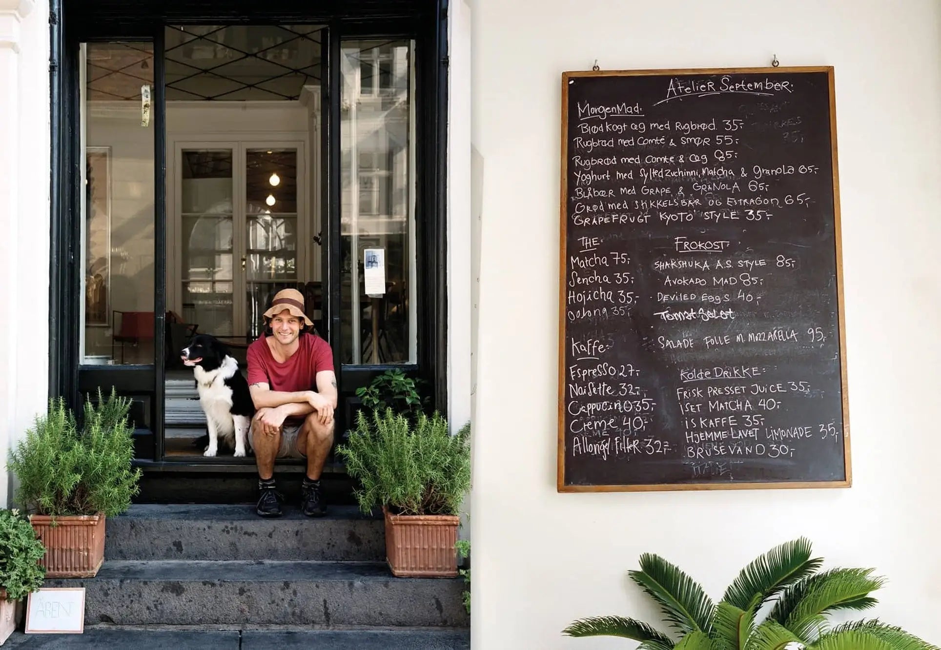 A man in a brown hat and red shirt sits on steps beside a black and white dog, smiling. Behind them is a glass door. To the right, an Apartamento Atelier September: A Place for Daytime Cooking chalkboard menu is visible.